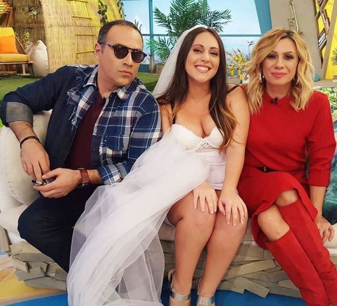 Η Κατερίνα από το Game of Love με νυφικό ανάμεσα σε δύο παρουσιαστές σε τηλεοπτικό πλατό.