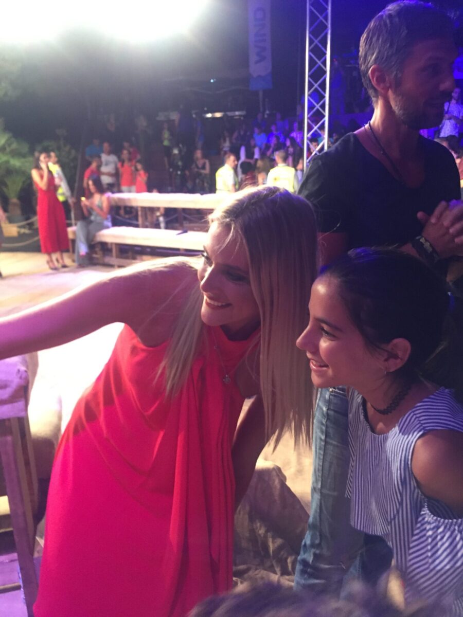 Μια ξανθιά γυναίκα με κόκκινο φόρεμα βγάζει σέλφι με ένα κορίτσι σε εκδήλωση.
