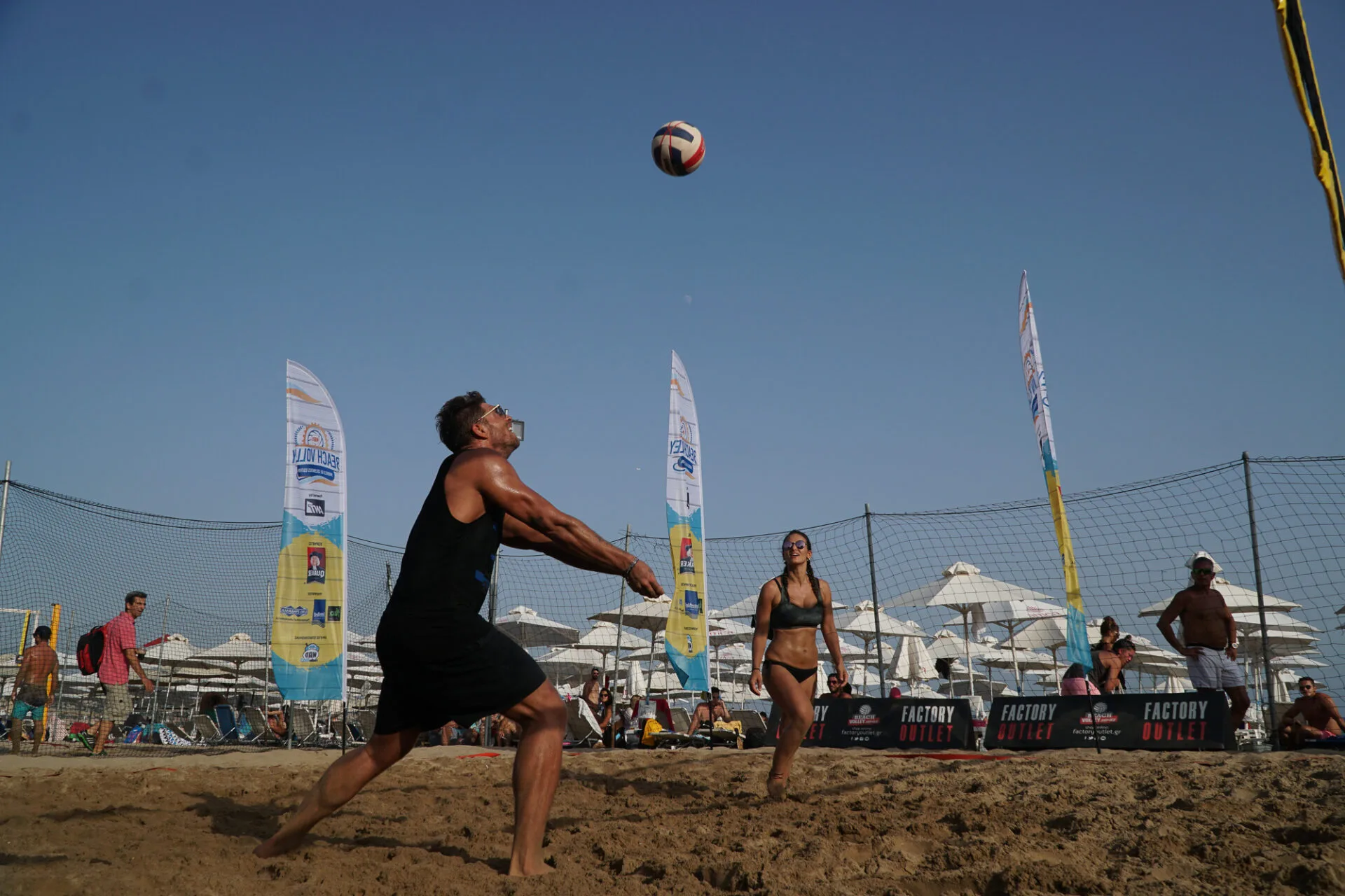 Άνδρας και γυναίκα παίζουν beach volley σε παραλία με κόσμο και ομπρέλες.