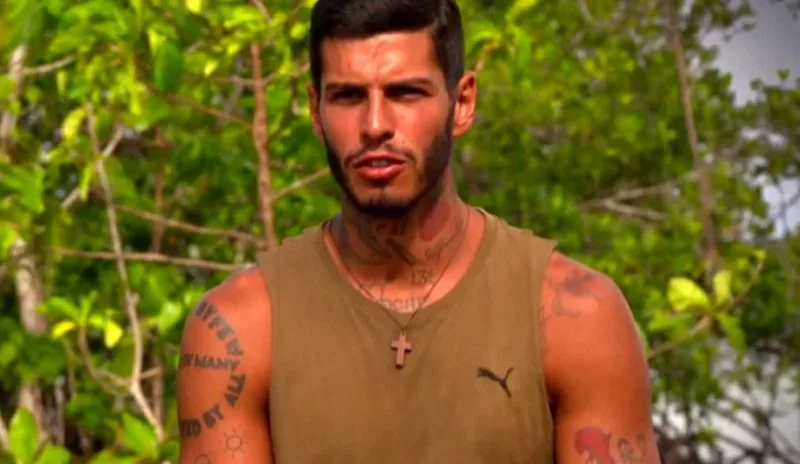 Παίκτης του Survivor 2 με τατουάζ και λαδί φανελάκι σε τροπικό φόντο.