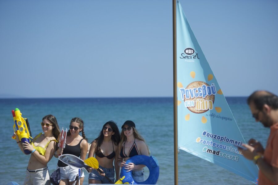 Τέσσερις κοπέλες στην παραλία με καλοκαιρινά αξεσουάρ δίπλα σε σημαία Mad Secret.