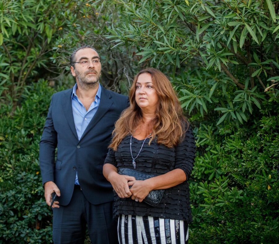 Ένας άνδρας με κοστούμι και μια γυναίκα με μαύρο τοπ και ριγέ παντελόνι σε εξωτερικό χώρο με πράσινο φόντο.
