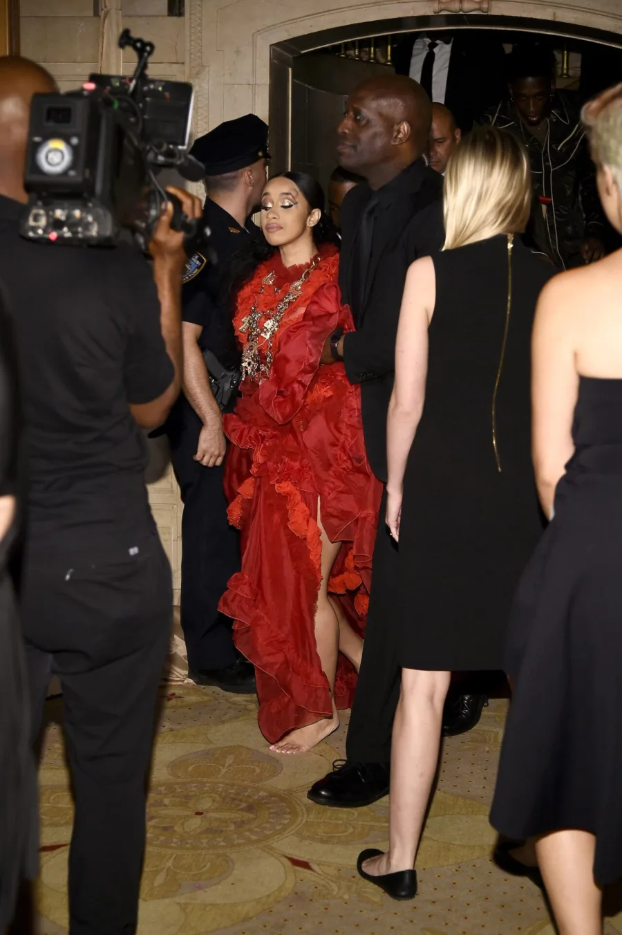Η Cardi B με κόκκινο φόρεμα και κοσμήματα, περιτριγυρισμένη από κόσμο και κάμερες.