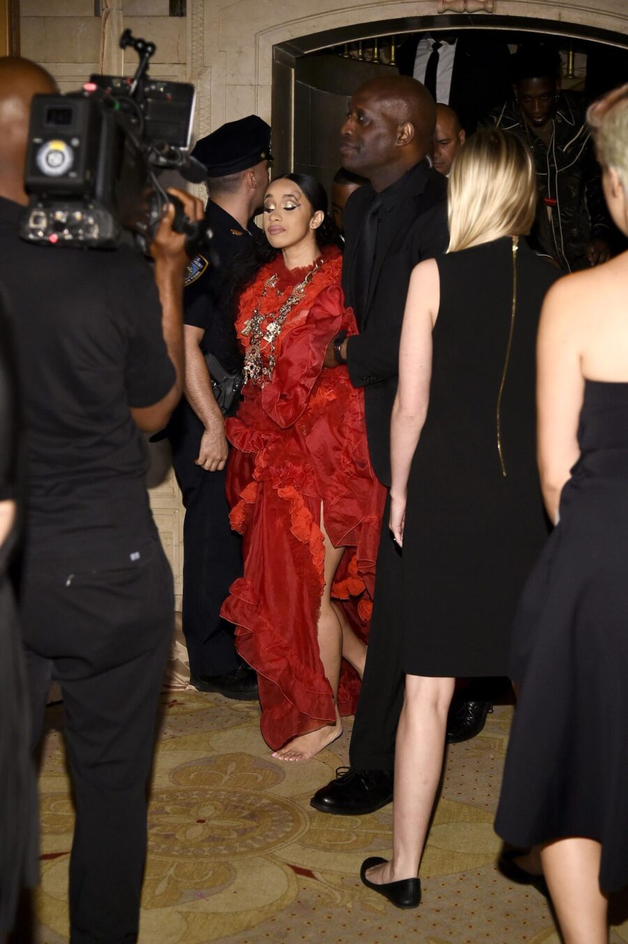 Η Cardi B με κόκκινο φόρεμα και κοσμήματα, περιτριγυρισμένη από κόσμο και κάμερες.