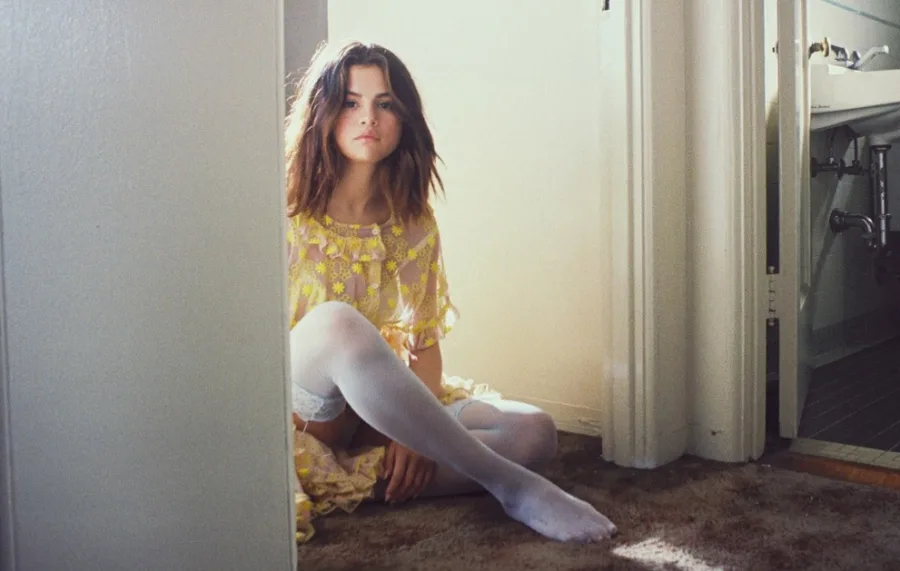 Η Selena Gomez κάθεται στο πάτωμα φορώντας κίτρινο δαντελωτό φόρεμα και λευκό καλσόν.