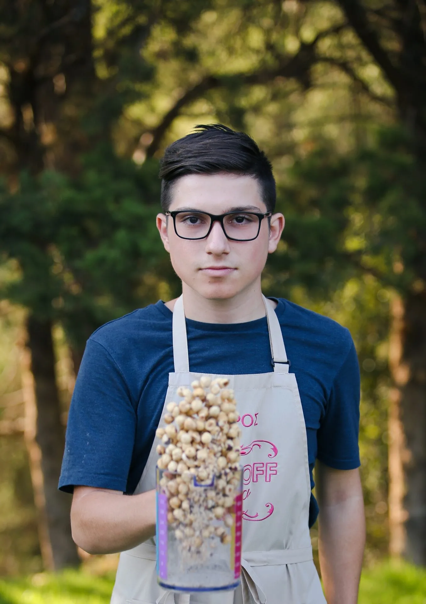 Νεαρός παίκτης του Bake Off Greece με ποδιά κρατάει ένα δοχείο με φουντούκια.