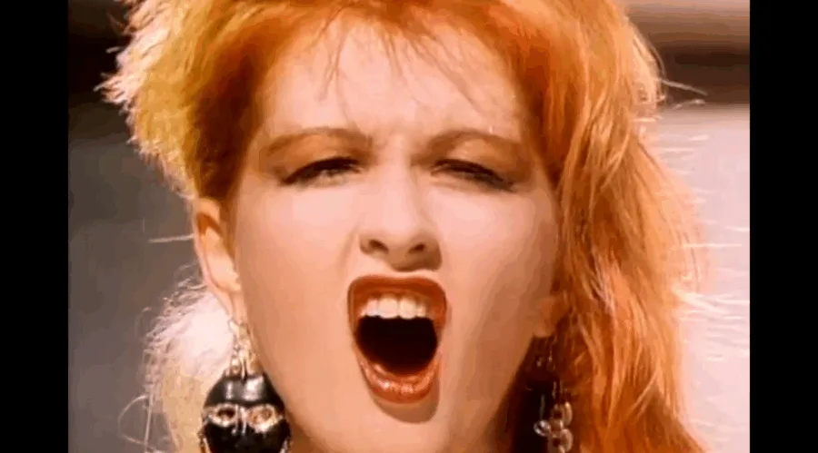 Κοντινό πλάνο της Cyndi Lauper με έντονο μακιγιάζ και πορτοκαλί μαλλιά.