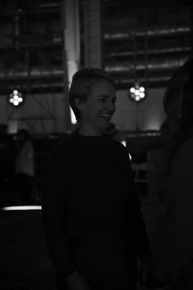 Γυναίκα με κοντό μαλλί χαμογελά σε backstage χώρο.