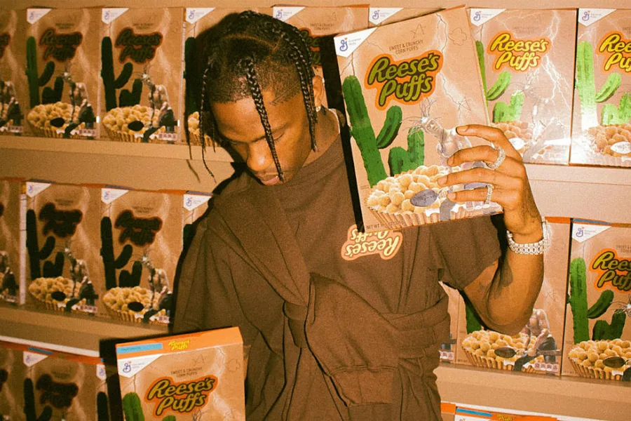 Ο Travis Scott κρατάει ένα συλλεκτικό κουτί δημητριακών Reese's Puffs μπροστά από ράφια γεμάτα με το ίδιο προϊόν.