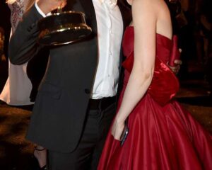 Ένας άνδρας με κοστούμι κρατά ένα βραβείο Emmy και αγκαλιάζει μια γυναίκα με κόκκινο φόρεμα.