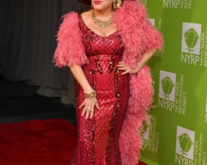 Η Bette Midler με εντυπωσιακό φούξια φόρεμα και καπέλο με φτερά σε κόκκινο χαλί.