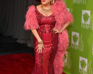 Η Bette Midler με εντυπωσιακό φούξια φόρεμα και καπέλο με φτερά σε κόκκινο χαλί.