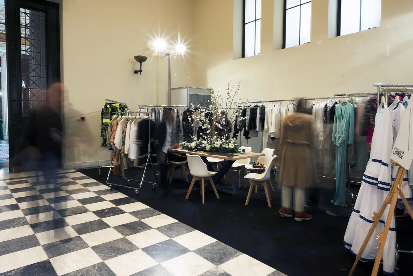 Εσωτερικός χώρος της έκθεσης Eleven The Fashion Project με κρεμάστρες ρούχων και τραπέζι.