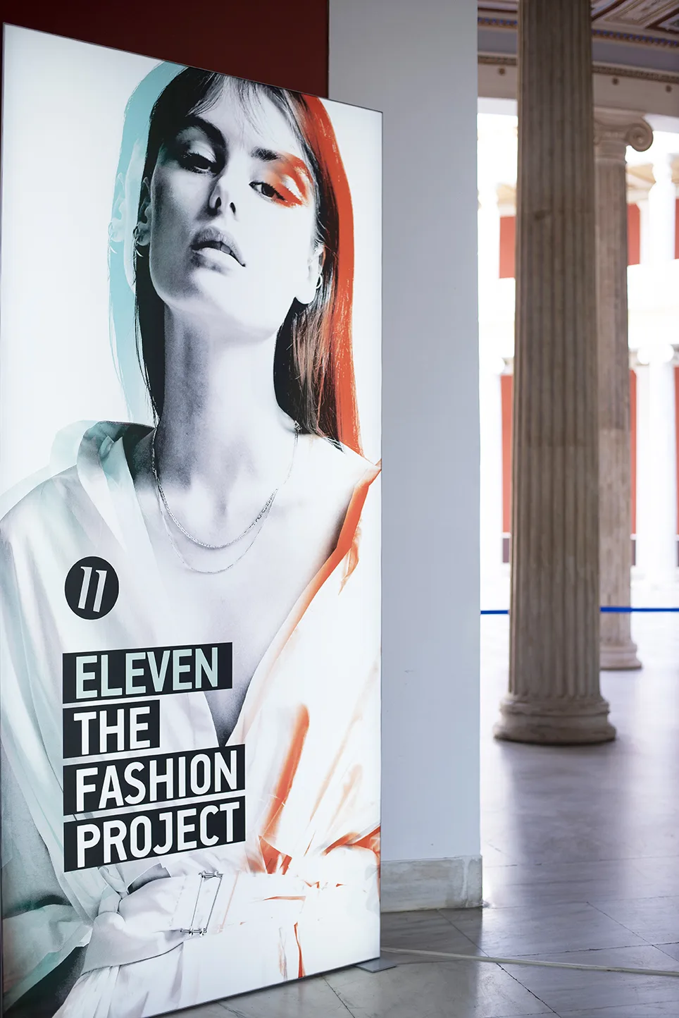 Αφίσα της έκθεσης Eleven The Fashion Project με γυναικείο πρόσωπο σε εσωτερικό χώρο με κίονες.