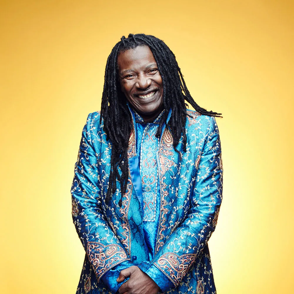 Ο Alpha Blondy χαμογελάει φορώντας μπλε παραδοσιακό ένδυμα σε κίτρινο φόντο.