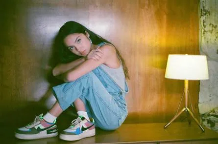 Η Olivia Rodrigo κάθεται σε ξύλινο πάγκο φορώντας τζιν σαλοπέτα και πολύχρωμα sneakers.