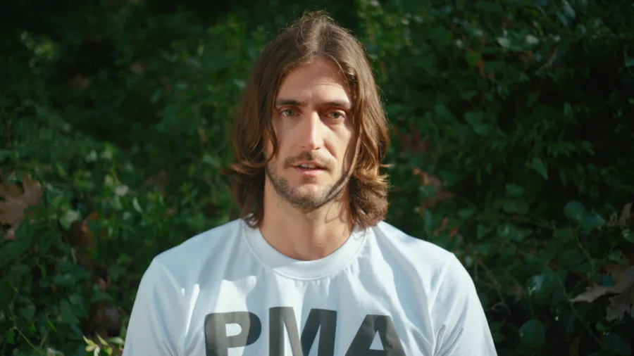 Ο rapper Ν.Ο.Ε. με λευκό t-shirt μπροστά από πράσινο φόντο με φυτά.