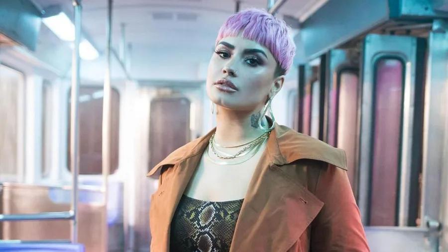 Η Demi Lovato με κοντά μωβ μαλλιά και καφέ παλτό μέσα σε βαγόνι τρένου.