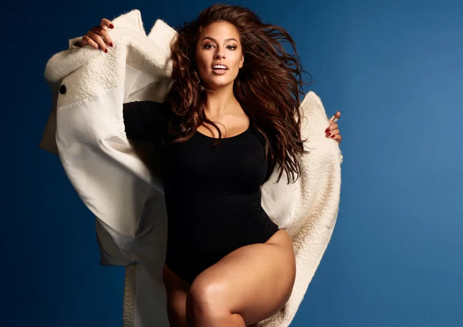 Η Ashley Graham ποζάρει με μαύρο κορμάκι και λευκό παλτό σε μπλε φόντο.