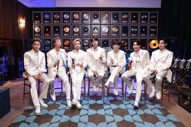 Το K-pop συγκρότημα BTS ποζάρει με λευκά κοστούμια σε δωμάτιο με χρυσούς δίσκους.