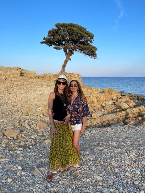 Η Δέσποινα Βανδή με φίλη της σε παραλία με βότσαλα και ένα δέντρο στο βάθος.