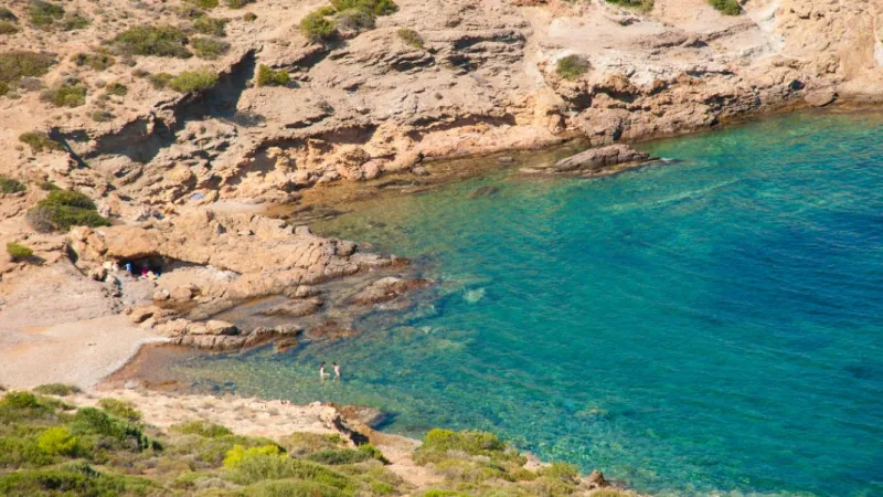 Πανοραμική άποψη μιας μικρής, απόμερης παραλίας με γαλαζοπράσινα νερά και βραχώδες τοπίο στην Αττική.