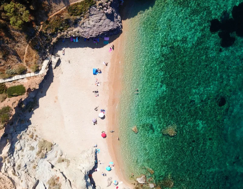 Αεροφωτογραφία από ψηλά μιας μικρής παραλίας με γαλαζοπράσινα νερά και λουόμενους στην Αττική.