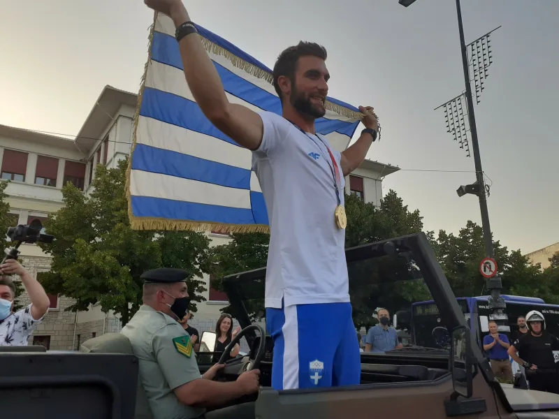 Ο Στέφανος Ντούσκος σε ανοιχτό όχημα κρατώντας την ελληνική σημαία.