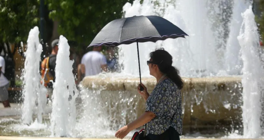 Γυναίκα με ομπρέλα μπροστά από σιντριβάνι σε ημέρα καύσωνα.
