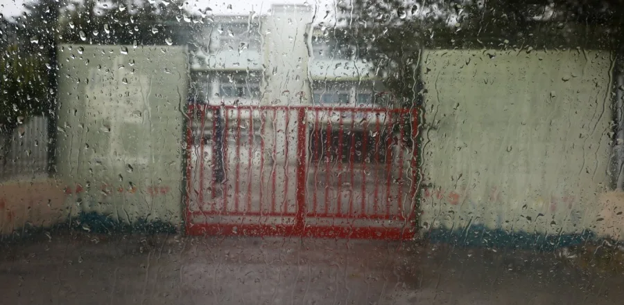 Σχολικό κτίριο με κόκκινη καγκελόπορτα μέσα από βρεγμένο τζάμι με σταγόνες βροχής.