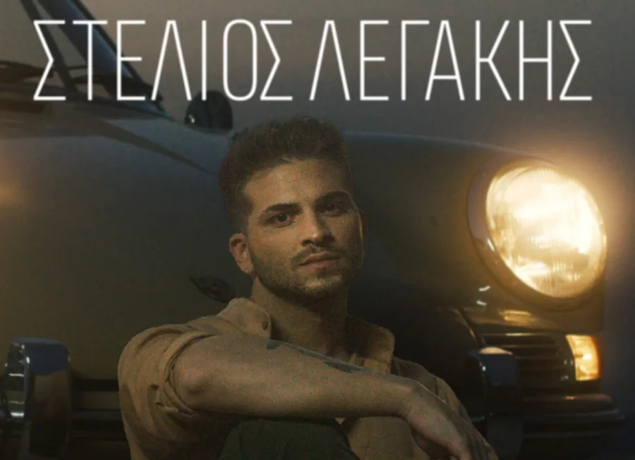 Ο Στέλιος Λεγάκης ποζάρει μπροστά από ένα κλασικό αυτοκίνητο με αναμμένο προβολέα.