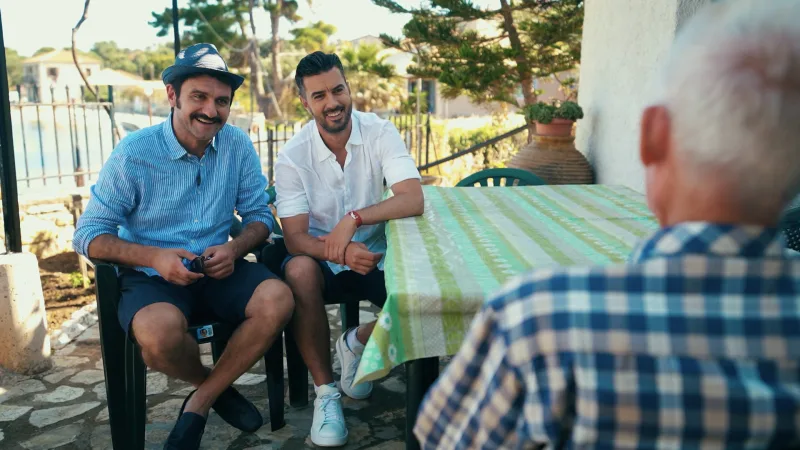 Ο Μελέτης Ηλίας και ο Γιάννης Τσιμιτσέλης κάθονται σε τραπέζι σε εξωτερικό χώρο.