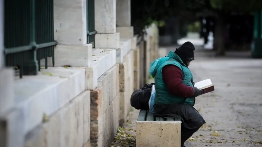 Άστεγος άνδρας κάθεται σε παγκάκι και διαβάζει βιβλίο σε εξωτερικό χώρο.
