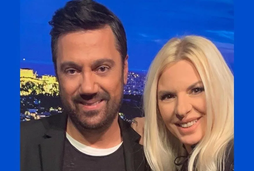 Ο Γιώργος Θεοφάνους και μια γυναίκα σε τηλεοπτικό πλατό με φόντο την Αθήνα.