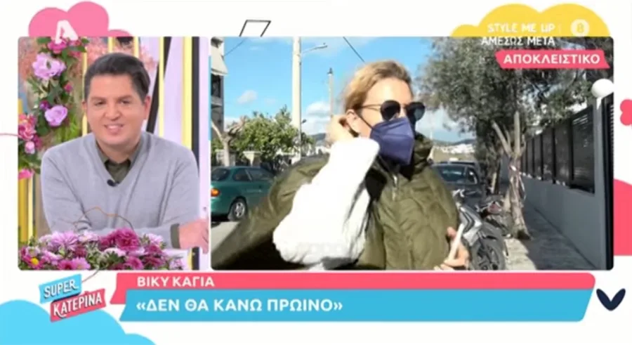 Η Βίκυ Καγιά με μάσκα προστασίας και λαδί μπουφάν σε εξωτερικό χώρο, δίπλα σε τηλεοπτικό πάνελ.