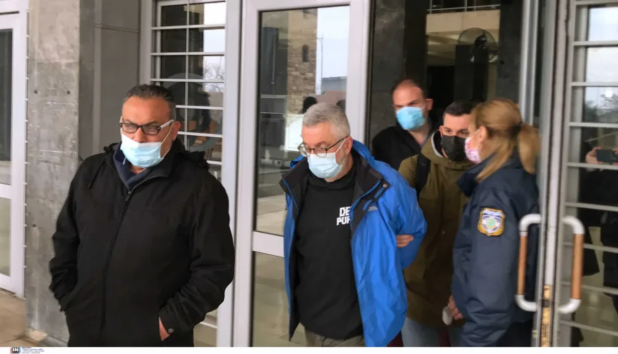 Ο Στάθης Παναγιωτόπουλος με μπλε μπουφάν και μάσκα προστασίας συνοδευόμενος από αστυνομικούς.