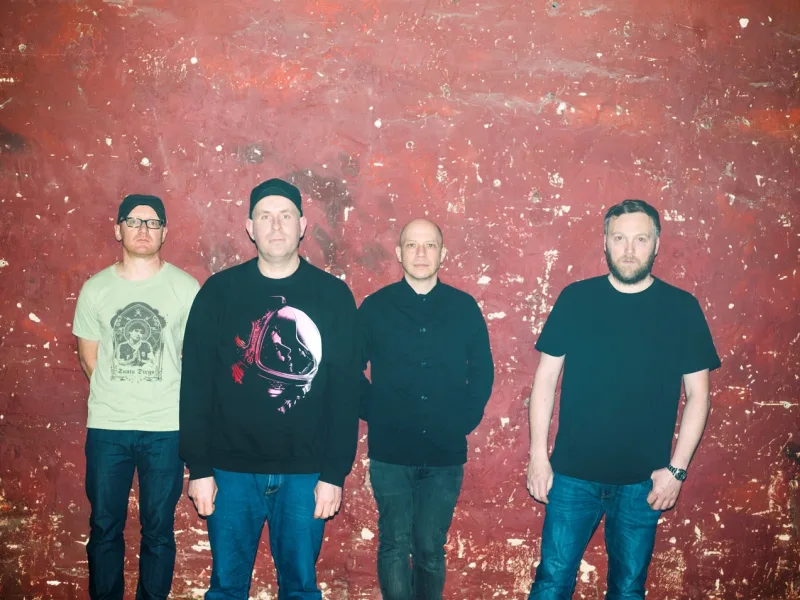 Το συγκρότημα Mogwai ποζάρει μπροστά από έναν κόκκινο τοίχο.