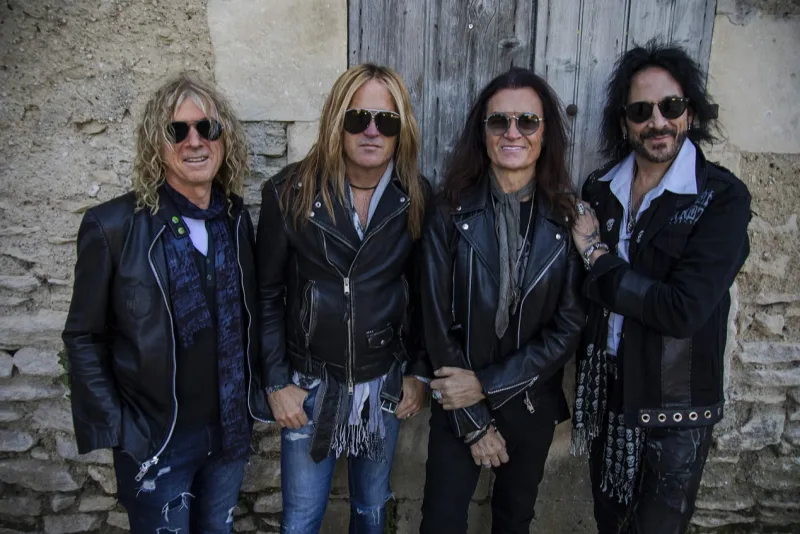 Τέσσερα μέλη του συγκροτήματος Judas Priest ποζάρουν μπροστά από πέτρινο τοίχο.
