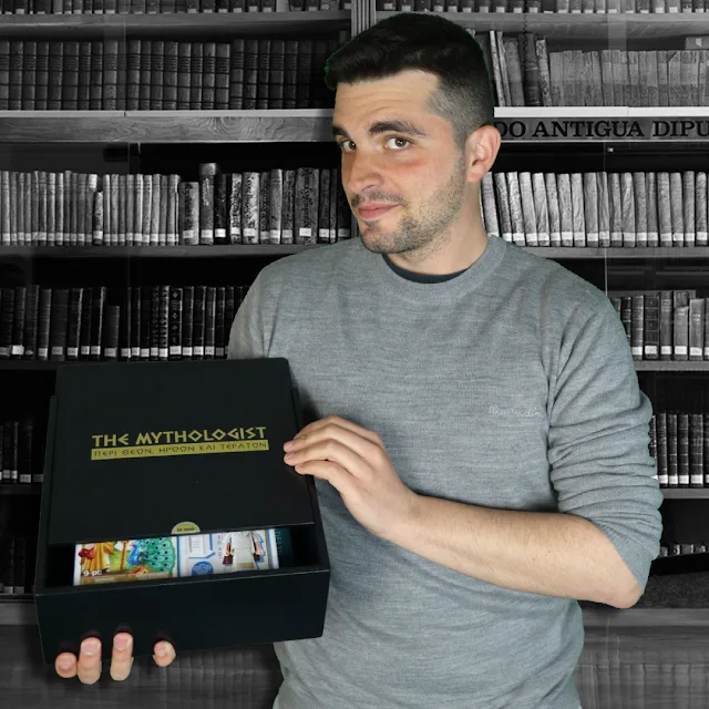 Ο The Mythologist κρατάει ένα μαύρο κουτί με το λογότυπό του μπροστά από μια βιβλιοθήκη.