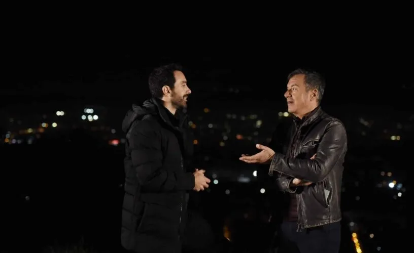 Ο Σάκης Τανιμανίδης συνομιλεί με έναν άνδρα σε εξωτερικό χώρο τη νύχτα με φόντο τα φώτα της πόλης.