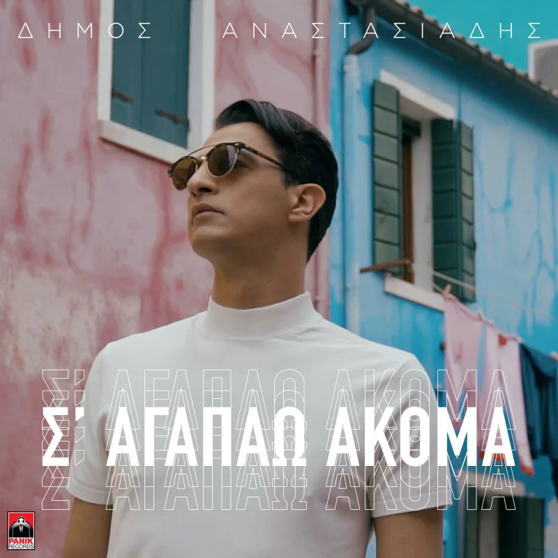 Ο Δήμος Αναστασιάδης με γυαλιά ηλίου και λευκό ζιβάγκο μπροστά από χρωματιστά κτίρια.