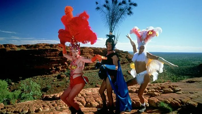 Τρεις drag performers με εντυπωσιακά κοστούμια και φτερά σε ερημικό τοπίο.