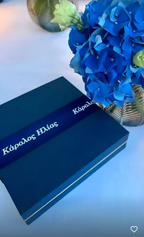 Μπλε κουτί με κορδέλα που γράφει Κάρολος Ηλίας δίπλα σε μπλε ορτανσίες.