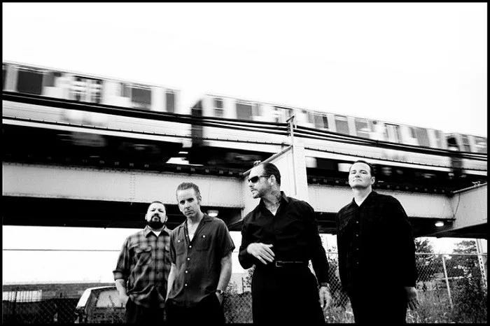 Ασπρόμαυρη φωτογραφία τετραμελούς rock συγκροτήματος μπροστά από γέφυρα με τρένο.
