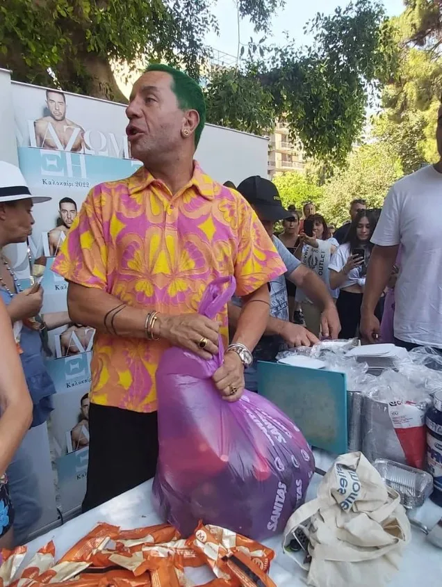 Ο Γιώργος Μαζωνάκης με πράσινα μαλλιά κρατάει μια μεγάλη μωβ σακούλα σε εξωτερικό χώρο.