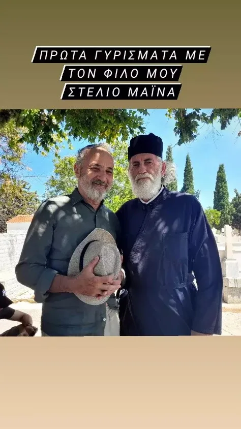 Ο Στέλιος Μάινας με έναν άνδρα ντυμένο ιερέα σε εξωτερικό χώρο.