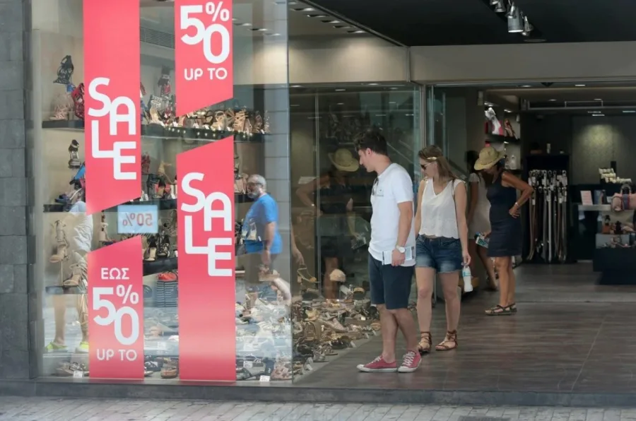 Βιτρίνα καταστήματος υποδημάτων με κόκκινα αυτοκόλλητα εκπτώσεων και κόσμο να κοιτάζει.