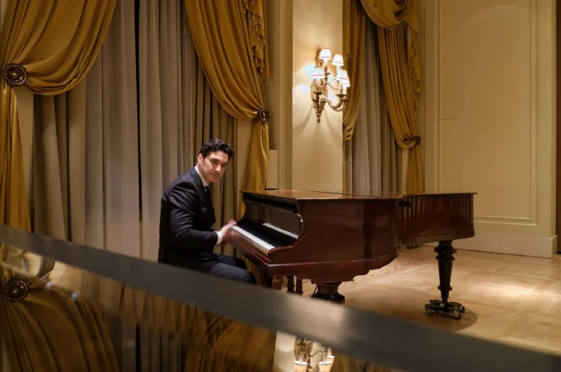 Ο Χριστόφορος Παπακαλιάτης ως Maestro παίζει πιάνο σε πολυτελές δωμάτιο.