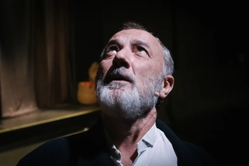 Κοντινό πλάνο άνδρα ηθοποιού με γκρίζα γενειάδα να κοιτάζει ψηλά σε θεατρική σκηνή.