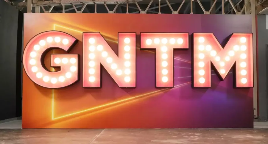 Το λογότυπο GNTM με φωτεινά γράμματα σε πορτοκαλί και μωβ φόντο.